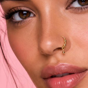 Puede incluir: Primer plano de un rostro con un piercing de nariz dorado. El piercing de nariz presenta un diseño curvo con una serie de pequeñas cuentas colgantes. La piel tiene un tono cálido y dorado, y el fondo es rosa claro.