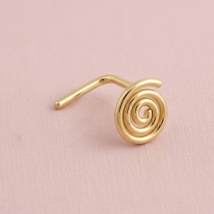 Op de afbeelding: Een goudkleurige neusring met een spiraalvormig ontwerp. De neusring heeft een gebogen staafje voor inbrengen en een opgerolde spiraal aan de voorkant. De sieraden staan tegen een zachtroze achtergrond.