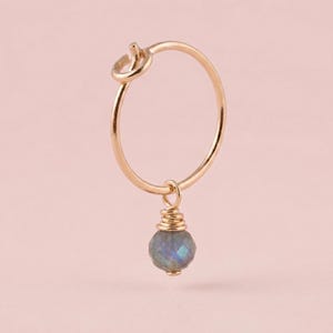 Peut inclure: Boucle d'oreille dorée avec un détail de nœud en haut. Une pierre précieuse bleu-gris à facettes est suspendue en bas de la boucle, enveloppée dans du fil d'or. La boucle d'oreille est posée sur un fond rose clair.