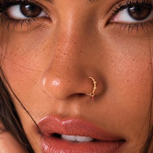 May include: A gold-toned nose ring hoop featuring three small metallic beads along the upper curve and a single dangling dark pink gemstone charm. The circular hoop is positioned in the nostril, showcasing a polished finish and delicate design.