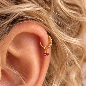 May include: A gold hoop earring featuring a series of small gold beads along the upper curve and two dangling, faceted, deep pink gemstone charms. The earring is secured through the cartilage of an ear, showcasing a polished metallic finish.