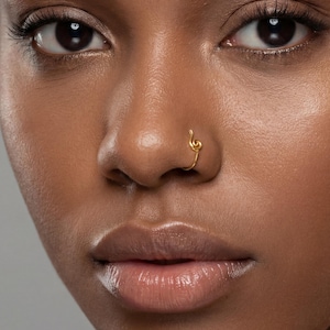 Może przedstawiać: Zbliżenie na twarz osoby, skupiając się na złotym kolczyku w nosie. Kolczyk w nosie ma kształt klucza wiolinowego. Odcień skóry jest ciepły brązowy, a usta błyszczące.