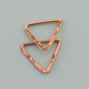 May include: Two hammered metal triangle earrings, one gold and one copper, are displayed against a light blue background. The earrings have a textured surface and a simple, geometric design. The triangles are slightly overlapping.