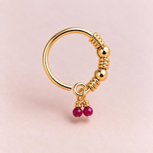 May include: A gold-toned hoop ring featuring a series of metallic beads and ribbed spacers along one side. Two small, round, deep pink faceted beads hang from a central loop attached to the hoop, creating a dangling charm effect on the jewelry.