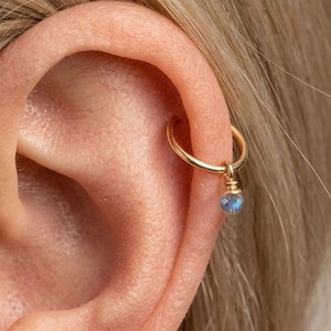 Peut inclure: Boucle d'oreille dorée avec une petite pierre précieuse bleue pendante. La boucle d'oreille est attachée à l'oreille. La pierre précieuse est facettée, captant la lumière. Ce bijou est de conception délicate.