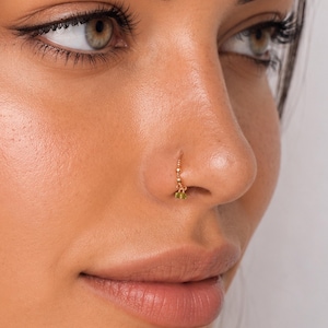 May include: A gold nose hoop ring featuring a series of small gold beads along the upper curve and a single, small, faceted light green gemstone charm dangling from the bottom of the hoop, securely positioned in the nostril piercing.