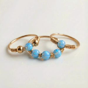 Puede incluir: Tres anillos dorados con cuentas de ópalo azul claro. Los anillos tienen un delicado diseño envuelto en alambre, con las cuentas de ópalo agrupadas en el centro de un anillo y como cuentas individuales en los otros dos. El fondo es blanco.