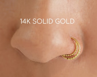 May include: A close-up of a nose with a 14K solid gold double hoop nose ring. The nose ring has a beaded design on the inner hoop and is a bright gold color.