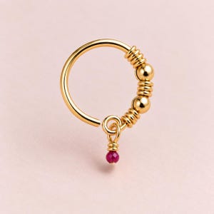 May include: A gold hoop ring featuring a series of smooth gold beads and ribbed gold spacers along one side. A small gold jump ring hangs from the hoop, suspending a single, faceted, round, deep pink gemstone bead at the bottom of the jewelry piece.