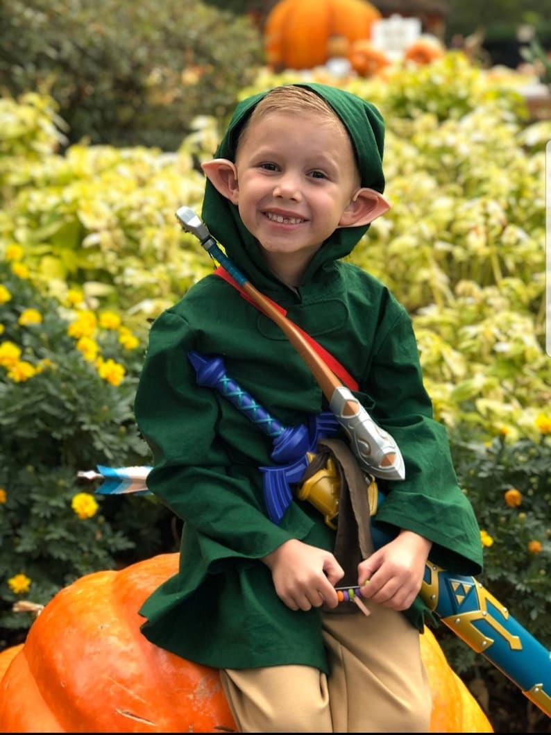 May include: A young boy dressed as Link from the Legend of Zelda video game series. He is wearing a green tunic, brown pants, and a pointy hat. He is holding a sword and a bow and arrow. He is sitting on a large orange pumpkin.
