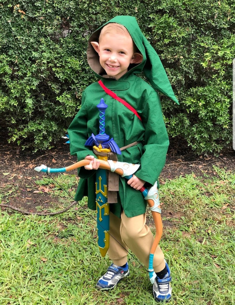 May include: A young child dressed as Link from the Legend of Zelda video game series. They are wearing a green tunic with a brown belt, a red sash, and a pointed hat with elf ears. They are holding a sword and a bow and arrow.