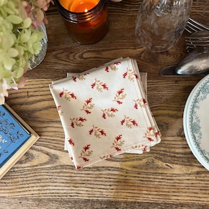 May include: A stack of folded cream-colored cloth napkins with a repeating pattern of red and gold oak leaves. The napkins are on a wooden table, alongside a lit candle, a glass, and a decorative plate.