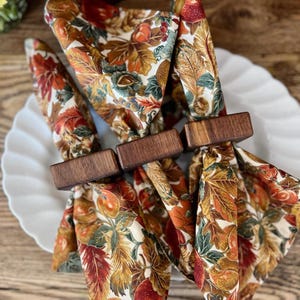 May include: A close-up of a white plate with three folded cloth napkins. The napkins are decorated with a fall leaf pattern in shades of orange, red, and green. Each napkin is held together by a rectangular wooden napkin ring.