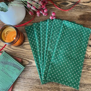 May include: A stack of six green cloth napkins with a white floral pattern. The napkins are arranged on a wooden surface, alongside a candle, a small book, and holiday decorations.