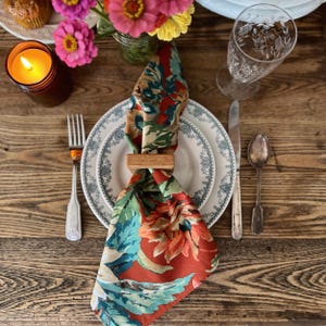 May include: A table setting with a floral napkin held by a wooden napkin ring, placed on a white plate with a blue floral border. A lit candle, silverware, and a glass of water are also on the wooden table.