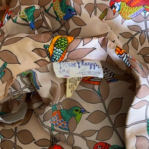 May include: A beige fabric with a repeating pattern of colorful birds and leaves. The fabric has a label that reads "Free Flaggs, A Div. of Edith Flaggs, California".