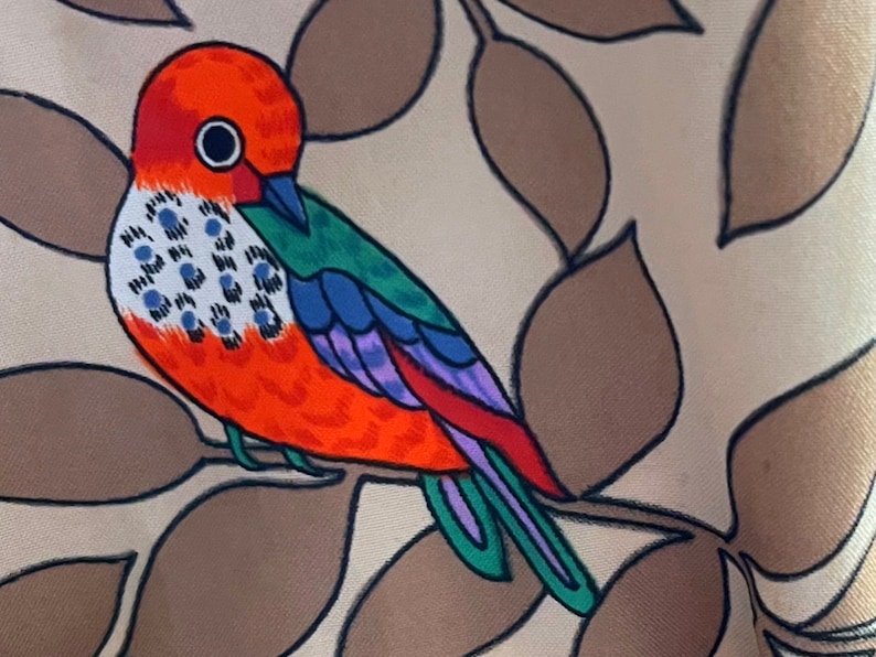 May include: A colorful bird with orange, red, green, blue, and purple feathers is perched on a branch with brown leaves. The bird has a large, round eye.