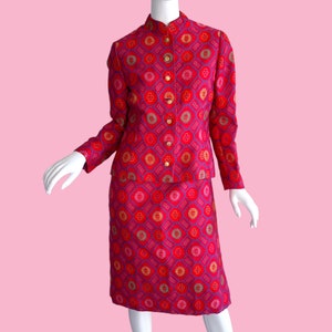May include: A vintage two-piece suit in a vibrant red hue, featuring a matching jacket and skirt. The jacket has a mandarin collar and gold buttons. The fabric is patterned with a geometric design in red, pink, and gold. The skirt is knee-length.