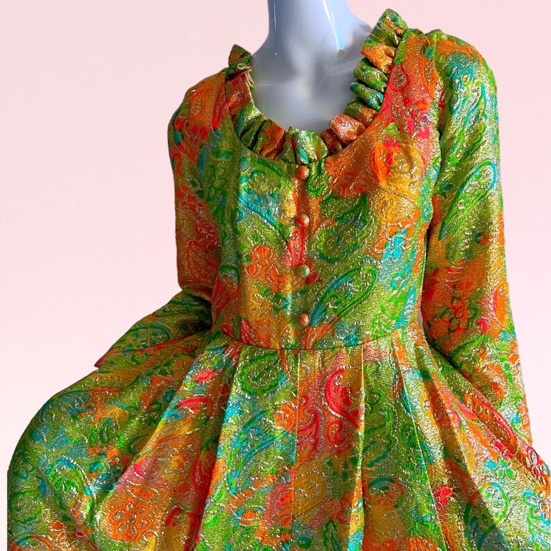 May include: A vintage dress with a green, orange, and pink paisley pattern. The dress has a ruffled neckline and long sleeves.