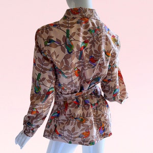 May include: A beige blouse with a long sleeve and a tie waist. The blouse has a colorful bird and leaf print.