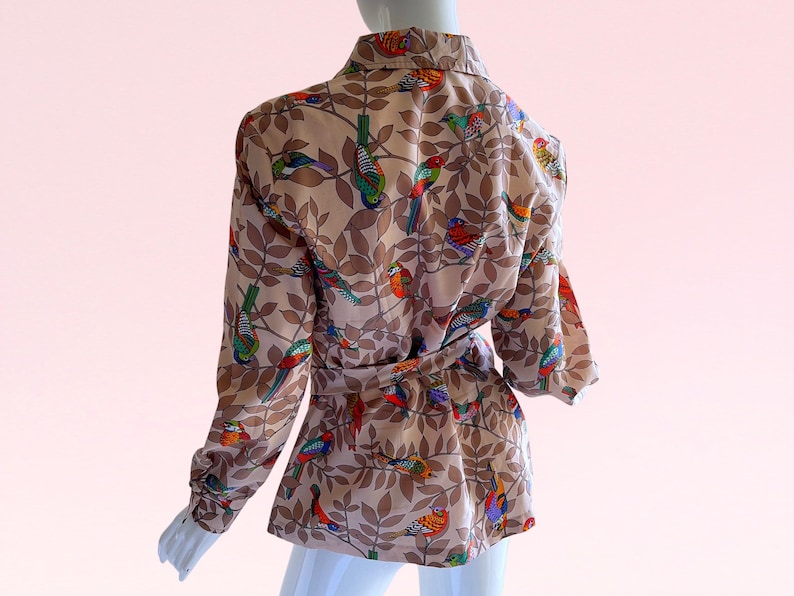 May include: A beige blouse with a long sleeve and a tie waist. The blouse has a colorful bird and leaf print.