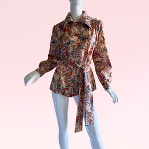May include: A beige long-sleeved shirt with a colorful bird and leaf print. The shirt has a tie belt and a collared neckline.