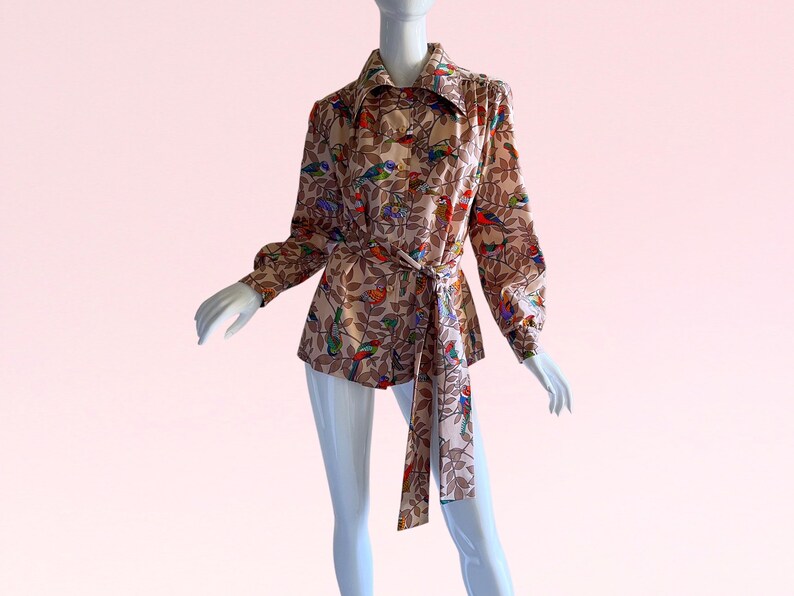 May include: A beige long-sleeved shirt with a colorful bird and leaf print. The shirt has a tie belt and a collared neckline.