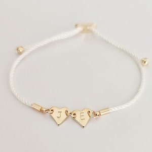 Puede incluir: Pulsera de cordón blanco con dos dijes de corazón de oro, cada uno con una letra grabada. Las letras son "J" y "E".