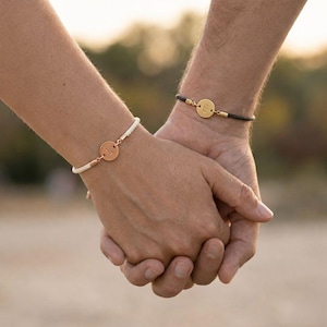 Puede incluir: Dos pulseras a juego con colgantes iniciales. Una pulsera tiene un cordón blanco y un colgante de oro rosa con la letra "L". La otra tiene un cordón negro y un colgante dorado con la letra "E". Las pulseras se llevan en las muñecas.