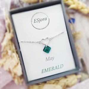 May include: A silver heart charm bracelet with a green emerald gemstone. The bracelet is in a white box with the text "Esjora" and "May Emerald".