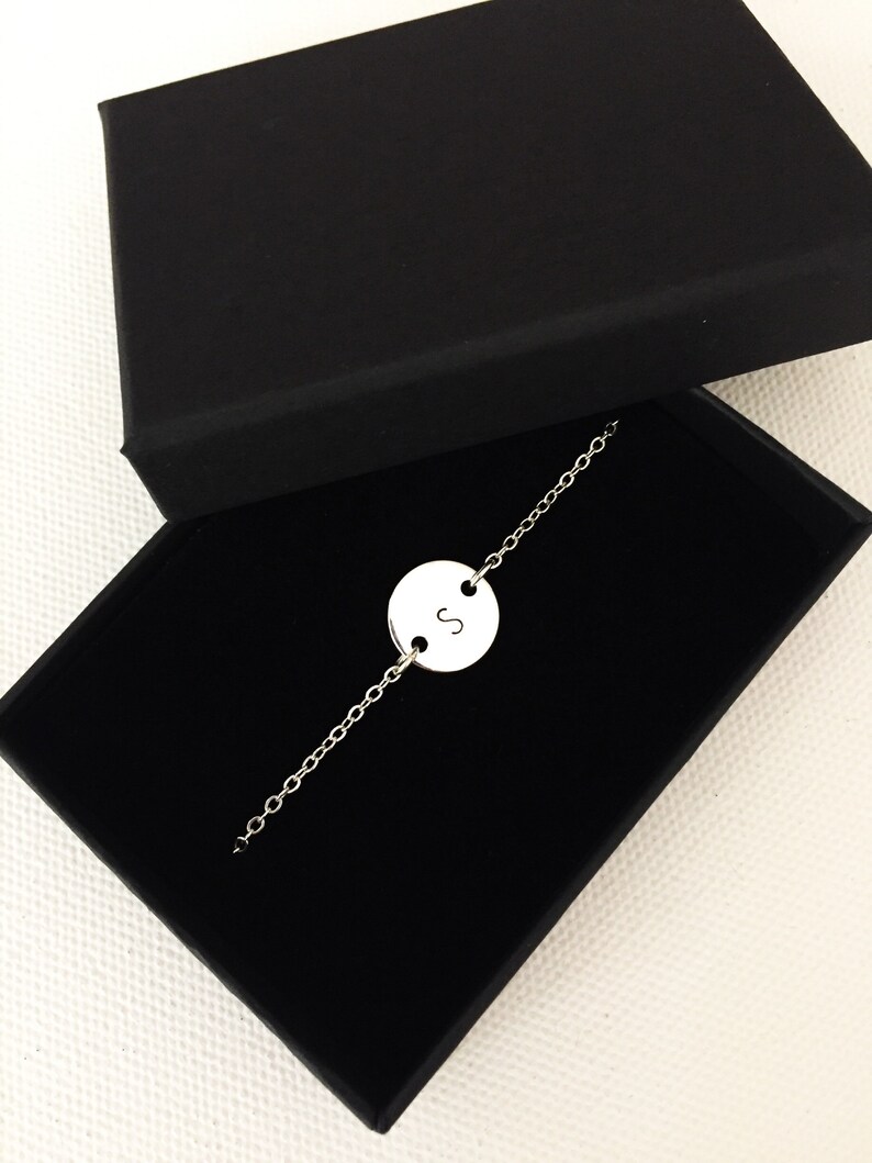 May include: A silver chain bracelet with a round charm featuring the letter "S" in a black box.