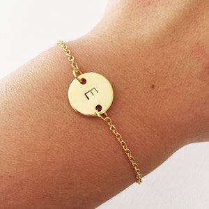 May include: A gold chain bracelet with a round charm featuring the letter "E" engraved on it.