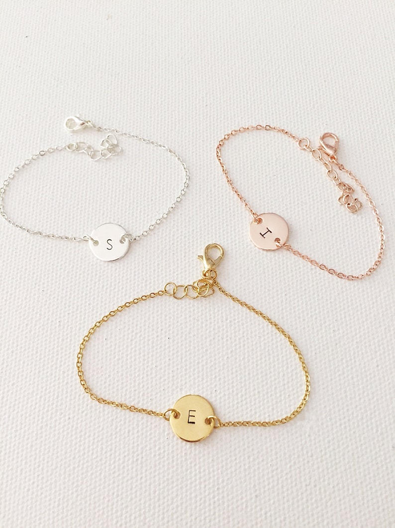 May include: Three delicate chain bracelets with a round charm featuring a single letter. The bracelets are silver, rose gold, and gold, and the charms are engraved with the letters S, I, and E.