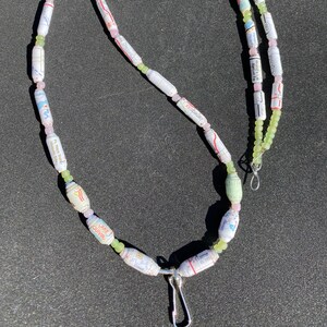 May include: A white, green, and pink beaded necklace made from rolled up paper with a silver clasp. The paper beads have text printed on them, including "Lake Quivira" and "Game".