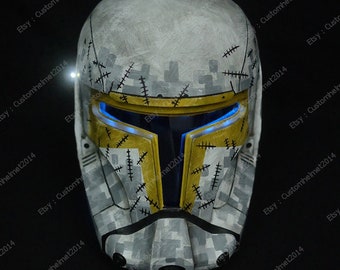May include: A white and gray camouflage helmet with yellow accents and a blue light. The helmet has a distressed look with stitching details.