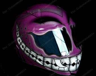 May include: A pink motorcycle helmet with a black and white graphic of a smiling face with sharp teeth. The helmet has a clear visor.