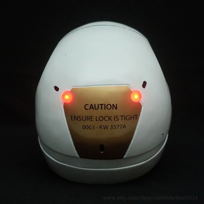 May include: White helmet with a gold plate that reads "CAUTION ENSURE LOCK IS TIGHT 0063-KW 3377A". The helmet has two red lights on the side.