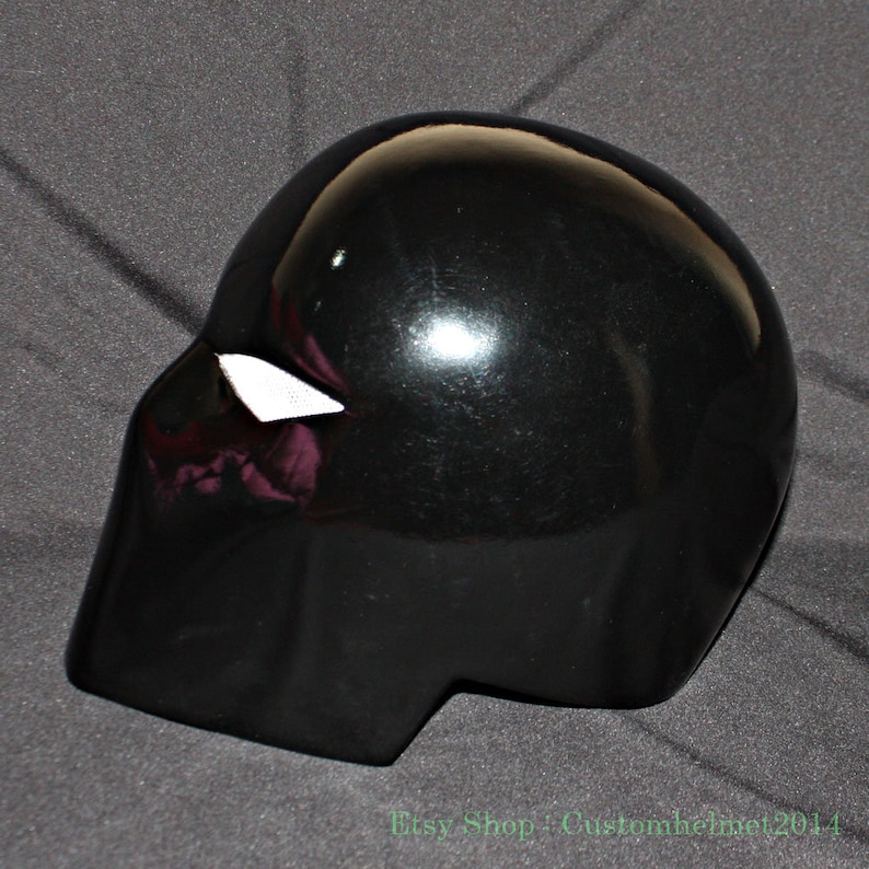 Pu&ograve; includere: Un casco nero lucido con una forma distintiva, caratterizzato da una piccola apertura triangolare bianca sul lato. La superficie del casco riflette la luce, evidenziando il suo design liscio e curvo. Il nome del negozio Etsy "Customhelmet2014" &egrave; visibile in basso.