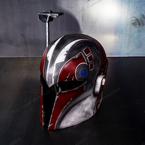 May include: A red, white, and gray helmet with a blue and red symbol on the side. The helmet has a black visor and a small antenna on top. The helmet is sitting on a gray surface.