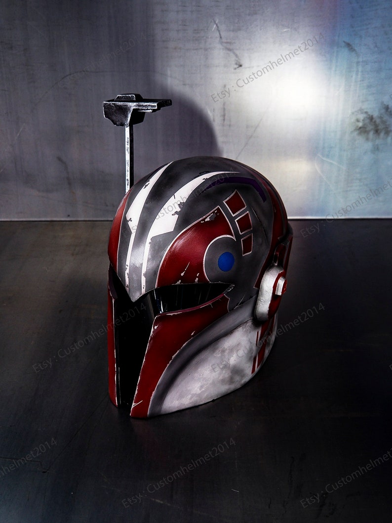 May include: A red, white, and gray helmet with a blue and red symbol on the side. The helmet has a black visor and a small antenna on top. The helmet is sitting on a gray surface.