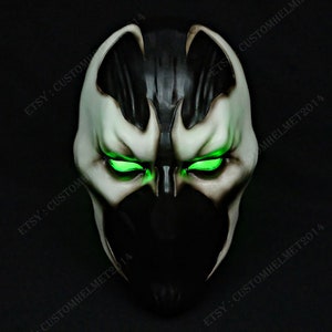 Puede incluir: Una máscara negra y blanca con ojos verdes brillantes. La máscara es una representación del personaje de cómic Spawn.