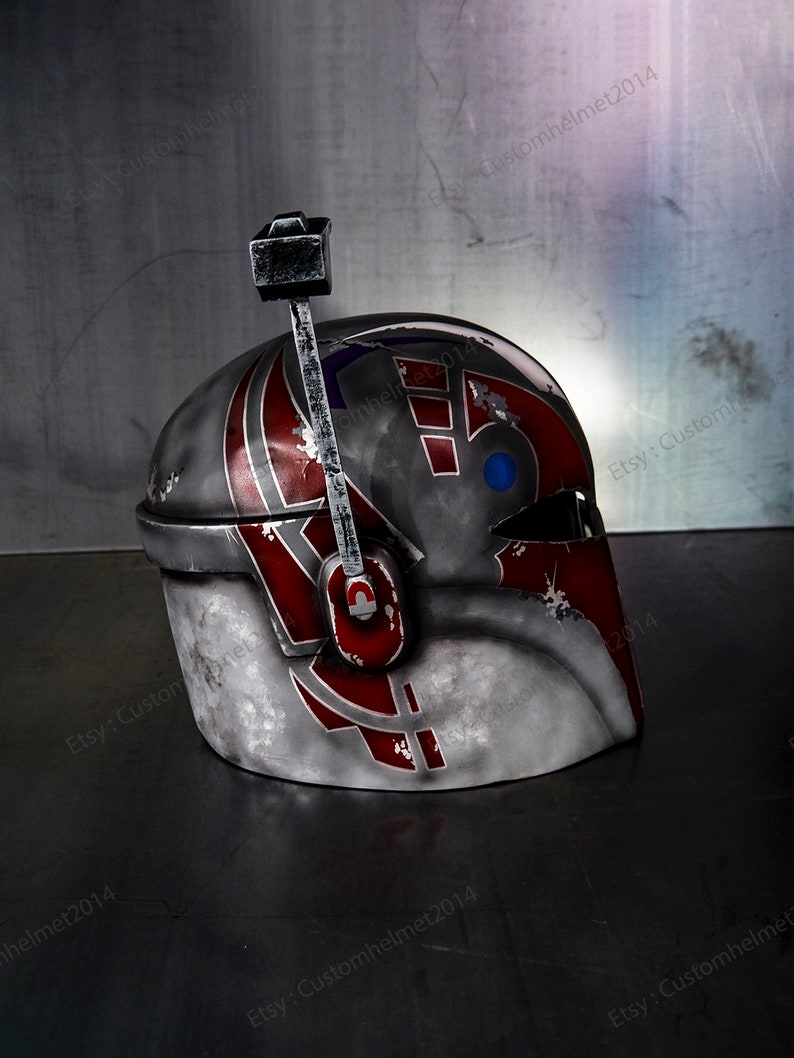 May include: A gray, red, and blue helmet with a weathered, distressed look. The helmet has a raised design on the side and a small, black antenna on top.