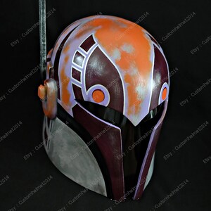 May include: A purple, orange, and gray Mandalorian helmet with a visor. The helmet has a weathered look with a design on the front.
