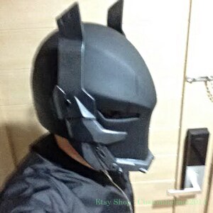 May include: A black and gray Batman helmet with a detailed design. The helmet has a visor and a textured surface.