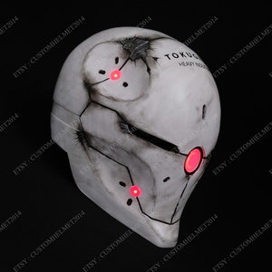 May include: A white helmet with a weathered, distressed look. The helmet has a cracked surface and a black visor. The helmet has red LED lights on the front and top. The text "TOKUGI HEAVY INDUS" is printed on the side of the helmet.
