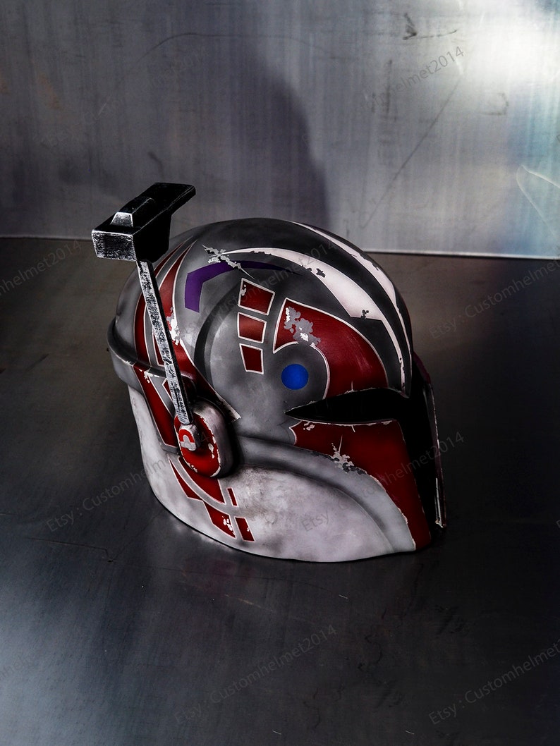 May include: A white, red, and blue helmet with a weathered finish. The helmet has a black visor and a black antenna on top. The helmet is sitting on a gray surface.