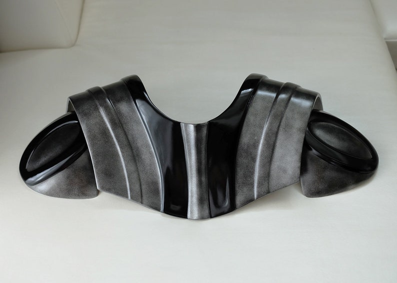 May include: A pair of black and silver shoulder armor pieces, designed to be worn around the neck and shoulders. The armor is made of a smooth, hard plastic and has a slightly textured surface.