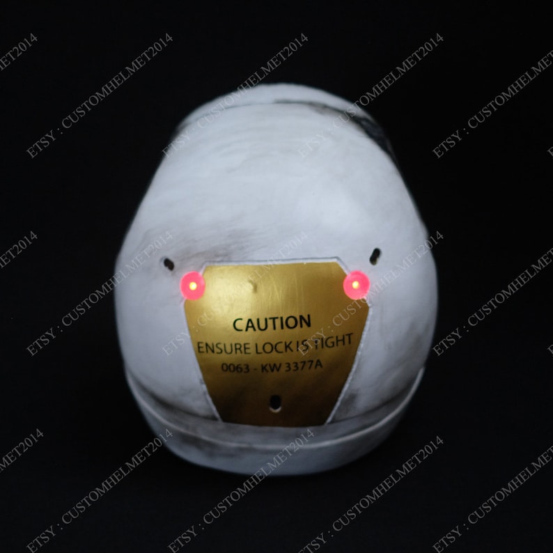 May include: A white helmet with a gold warning sign that reads "CAUTION ENSURE LOCK IS FIGHT 0063-KW 3377A". The helmet has two red lights on the front.