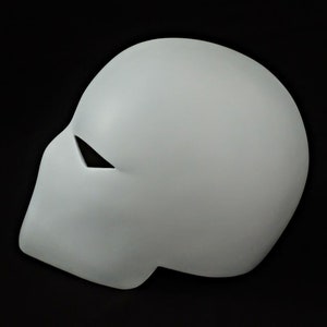 Pu&ograve; includere: Una maschera grigio chiaro con una superficie liscia e una singola apertura triangolare per gli occhi. La maschera &egrave; una forma solida e curva, probabilmente per cosplay o costumi, su sfondo nero.