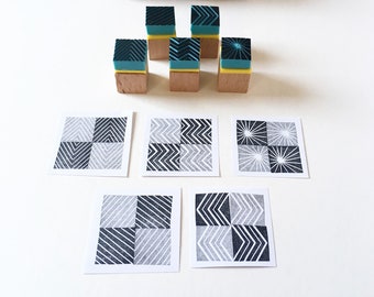 May include: Six wooden stamps with geometric designs in black and teal on a white background. The stamps are arranged in two rows of three. The stamps are packaged in a brown cardboard tube with a black and white geometric design. The stamps are used to create patterns on paper.